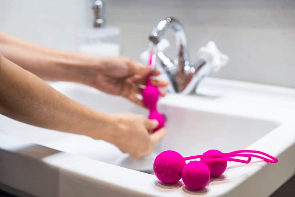 Hands washing pink silicone Ben Wa balls under a faucet.